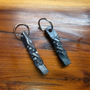 May include: Two hand-forged iron keychains featuring a square-profile bar with a central twisted design. Each piece has a dark, textured metallic finish and is attached to a circular steel split ring for holding keys securely.