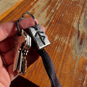 May include: A silver-toned metal keychain featuring a rectangular pendant with a dark, etched rune symbol. The pendant is attached to a circular metal ring holding several standard brass-colored keys and a black fabric strap extension.