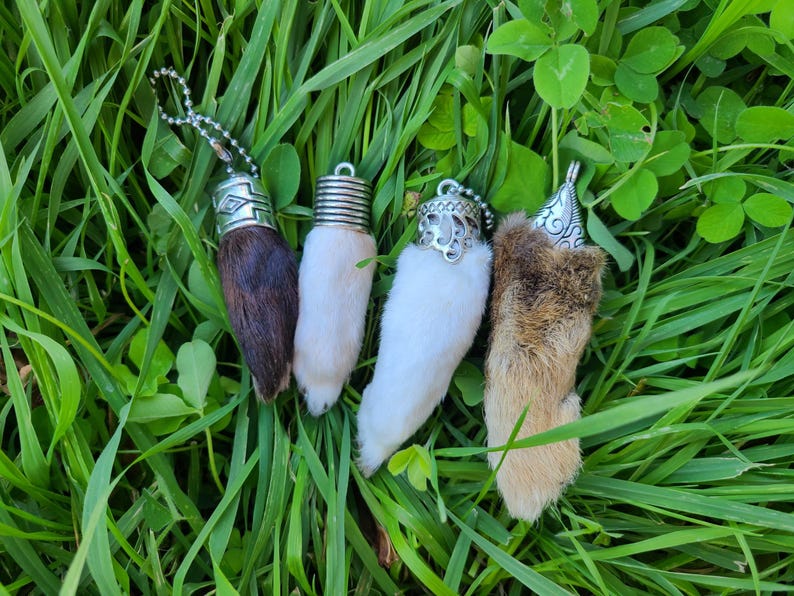 May include: Four different animal paw charms with silver metal accents. The charms are made of real fur and are attached to silver chains. The charms are displayed on a bed of green grass.