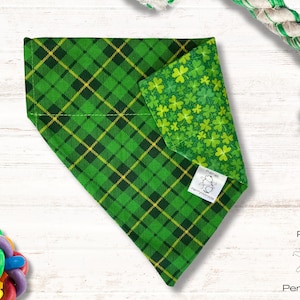 May include: Green plaid dog bandana with a shamrock print on the reverse side. The bandana is folded and has a small white tag with the text "Planet PerryWinkle" on it.