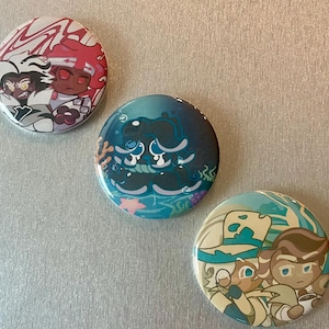 May include: Four round pinback buttons with colorful cartoon characters. The first button features a black and white character with red hair and a white background. The second button features a blue and white character with a blue background. The third button features a blue and white character with a blue background. The fourth button features a blue and white character with a blue background.