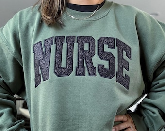 May include: An olive green sweatshirt with the word "NURSE" in large, glittery, navy blue letters across the chest. The sweatshirt has a crew neck and long sleeves. The overall style is casual and comfortable.
