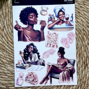 May include: A sheet of stickers featuring illustrations of Black women in various self-care activities, including taking a bath, reading a book, and applying makeup. The stickers are pink and white and have the text "me time stickers" on the top.