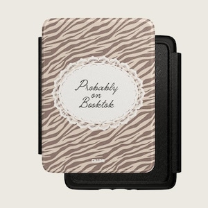 Può includere: Una custodia nera per e-reader con un motivo a strisce zebrate beige e marrone. Al centro della parte anteriore si trova un'etichetta ovale bianca bordata di pizzo con il testo nero "Probably on Booktok" e la parola "CHARM" in basso.