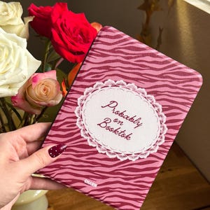 May include: A rectangular e-reader case featuring a pink and dark red zebra stripe pattern. The center displays a white circular lace-trimmed emblem with the text "Probably on Booktok" in a dark red script font and the word "CHARN" at the bottom.