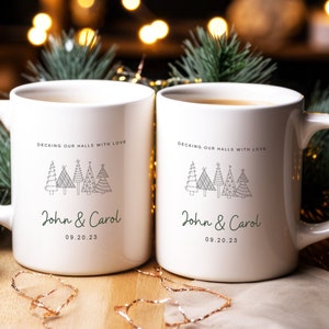 May include: Two white ceramic mugs with black handles. Each mug has a design of stylized Christmas trees and the text "DECKING OUR HALLS WITH LOVE" and "John & Carol" with the date "09.20.23".
