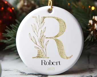 Puede incluir: Adorno navideño de cerámica blanca con una letra R dorada y un diseño floral dorado. El adorno está personalizado con el nombre "Robert" y el año "2023".