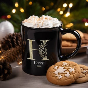 May include: Black ceramic mug with a gold letter H and a gold leaf design. The mug is filled with a hot beverage topped with whipped cream. The mug has the name "Henry" and the year "2023" printed on it.