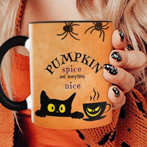 Puede incluir: Taza de cerámica naranja con asa negra. La taza tiene un gato negro asomándose por la parte inferior, una taza humeante de café con una cara espeluznante y el texto "Pumpkin spice and everything nice" en negro con un corazón morado.