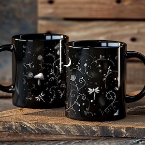 May include: Two black ceramic mugs with white floral and celestial designs. One mug features a mushroom, the other a crescent moon. The mugs are on a wooden surface.