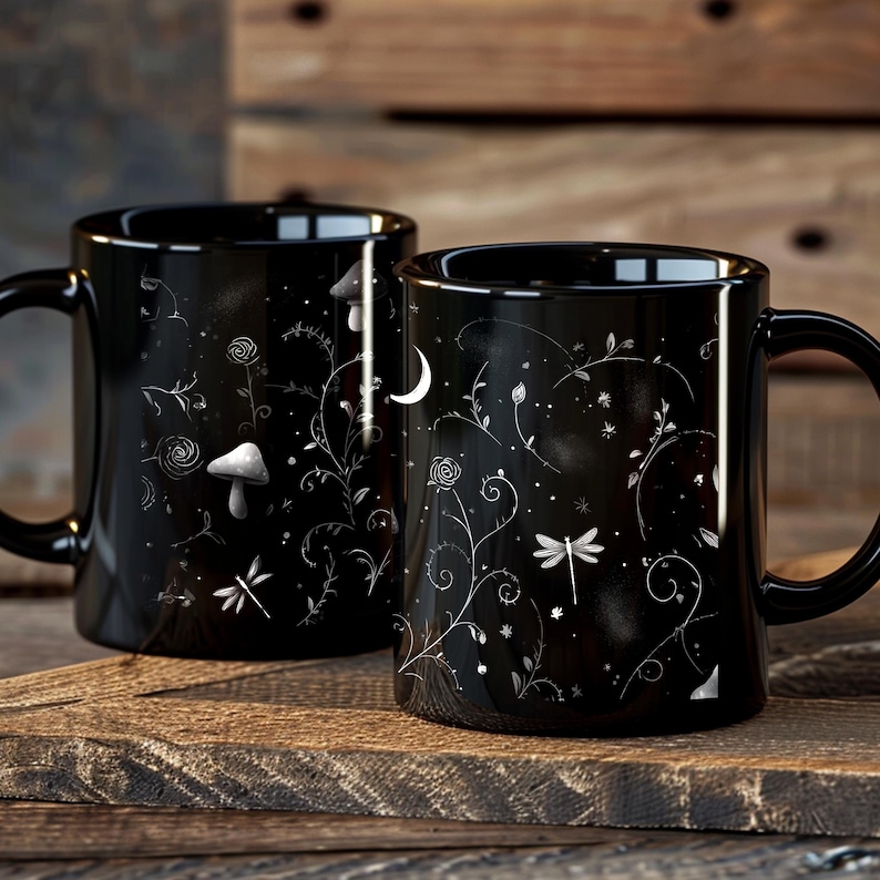 May include: Two black ceramic mugs with white floral and celestial designs. One mug features a mushroom, the other a crescent moon. The mugs are on a wooden surface.