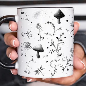 May include: White ceramic mug with a black rim and handle, featuring a whimsical black and white botanical design. The design includes mushrooms, flowers, and dragonflies. The mug is held by a person.