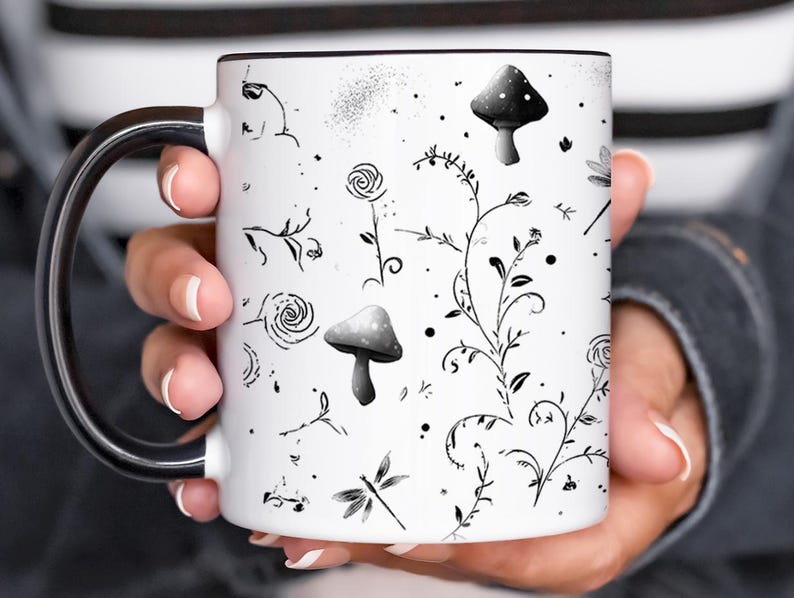 May include: White ceramic mug with a black rim and handle, featuring a whimsical black and white botanical design. The design includes mushrooms, flowers, and dragonflies. The mug is held by a person.
