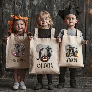 Peut inclure: Trois enfants d&eacute;guis&eacute;s pour Halloween tiennent des sacs de bonbons personnalis&eacute;s. Les sacs sont en toile et repr&eacute;sentent une sorci&egrave;re, une citrouille et une toile d'araign&eacute;e. Les sacs sont personnalis&eacute;s avec les noms "Olivia", "Adam" et "Penelope".