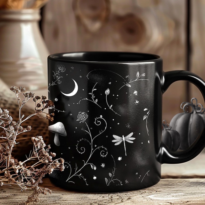May include: Black ceramic mug with a whimsical design. The mug features white illustrations of a crescent moon, mushroom, dragonfly, flowers, and stars. The handle is shaped like a pumpkin. The mug is set against a rustic wooden background.