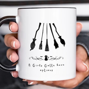 May include: White ceramic mug with a black rim and handle, featuring a design of five black witch brooms. The mug has the text "A Girls Gotta have options" with a small black cat and decorative elements, perfect for Halloween.