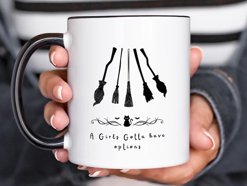 May include: White ceramic mug with a black rim and handle, featuring a design of five black witch brooms. The mug has the text "A Girls Gotta have options" with a small black cat and decorative elements, perfect for Halloween.