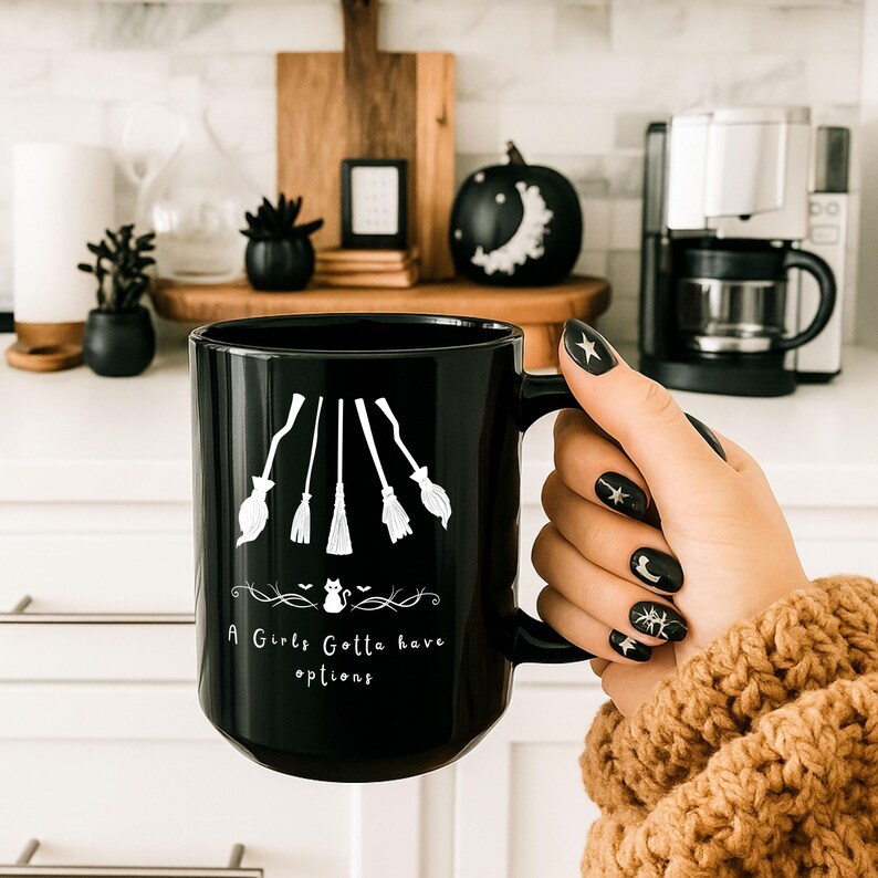 May include: Black ceramic mug with white witch broom graphics and the text "A Girls Gotta have options". The mug has a large handle and is held by a person with black nail polish with star and moon designs. The mug is a Halloween-themed product.