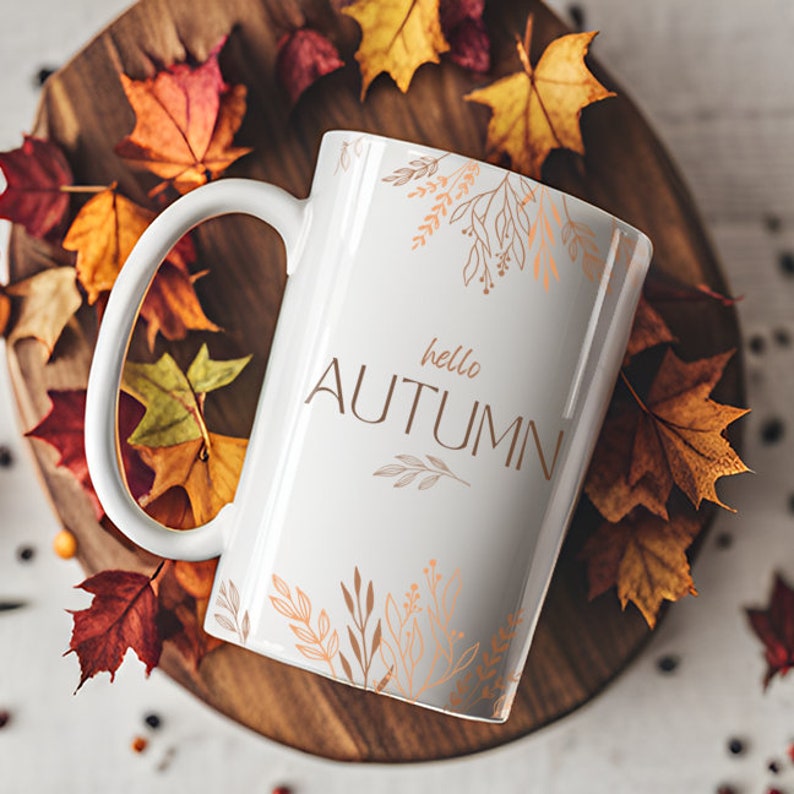 May include: White ceramic mug with a gold outline of leaves and the text "hello autumn" in gold lettering. The mug is surrounded by fall leaves on a wooden surface.