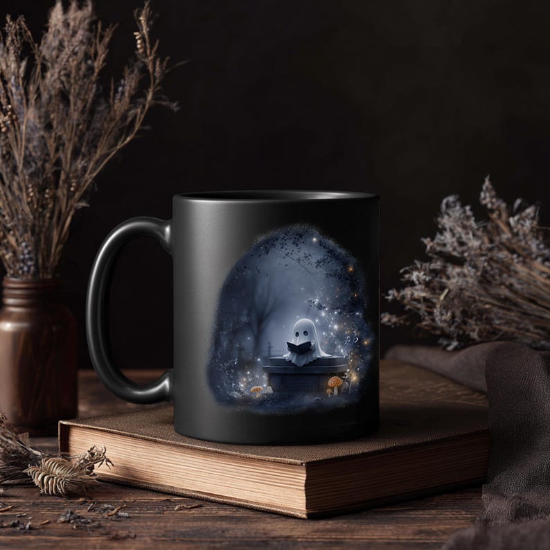 Puede incluir: Taza de cer&aacute;mica negra con una ilustraci&oacute;n caprichosa de un fantasma leyendo un libro en un bosque espeluznante. El dise&ntilde;o incluye un fondo azul oscuro con estrellas y setas brillantes. La taza est&aacute; sobre un libro marr&oacute;n.