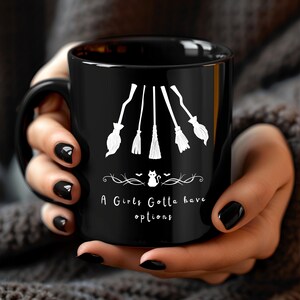 May include: Black ceramic mug featuring a white graphic of five witch brooms. Below the brooms is a decorative line with a cat and bats. The text "A Girls Gotta have options" is printed below the graphic.
