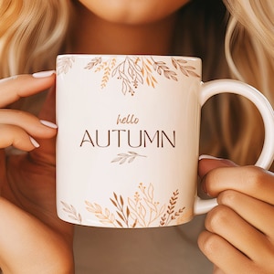 May include: White ceramic mug with a brown outline of leaves and branches around the top and bottom. The text "hello autumn" is printed in brown on the mug.
