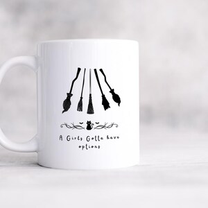 May include: White ceramic mug with a black graphic of five witch brooms and the text "A Girls Gotta have options". The brooms are arranged in a descending order. A decorative line with a cat and bats is below the text.