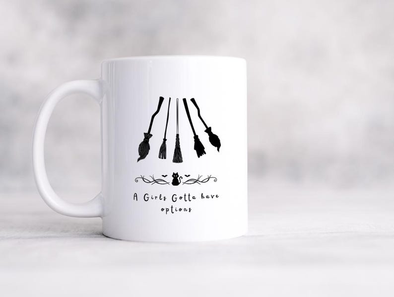 May include: White ceramic mug with a black graphic of five witch brooms and the text "A Girls Gotta have options". The brooms are arranged in a descending order. A decorative line with a cat and bats is below the text.