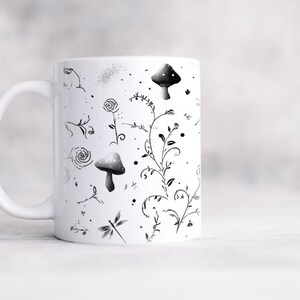 May include: White ceramic mug featuring a black and gray botanical design. The design includes mushrooms, flowers, vines, and a dragonfly. The mug has a curved handle and a smooth, glossy finish.