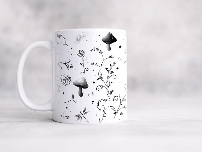 May include: White ceramic mug featuring a black and gray botanical design. The design includes mushrooms, flowers, vines, and a dragonfly. The mug has a curved handle and a smooth, glossy finish.