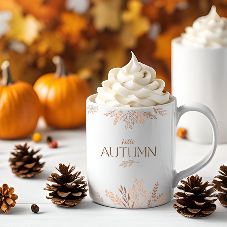 May include: White ceramic mug with a brown and orange floral design and the text "hello autumn". The mug is filled with a white whipped cream topping. The mug is sitting on a white surface with pine cones and fall leaves in the background.