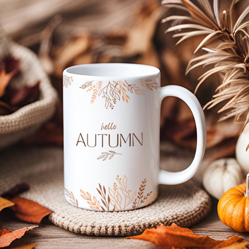 May include: White ceramic mug with a brown and orange floral design and the text "hello autumn". The mug is sitting on a knitted surface with fall leaves scattered around it.
