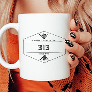 Puede incluir: Taza de café de cerámica blanca con un diseño gráfico en blanco y negro. El diseño presenta una forma hexagonal con el texto "Creepin' it real in the 313 since 1988". El gráfico también incluye dos manos de esqueleto haciendo un signo shaka.