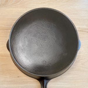 May include: A black cast iron skillet with a long handle and a round cooking surface. The skillet has the text "WAGNER" and "SIDNEY" embossed on the surface, along with the number "9". This vintage cookware is ideal for cooking.