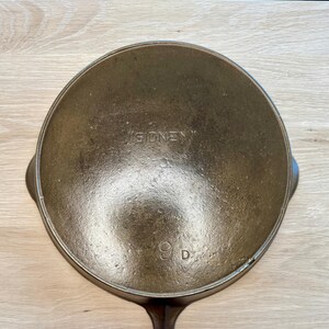 May include: A vintage cast iron skillet with a long handle and a circular cooking surface. The word "SIDNEY" is embossed on the bottom, along with the number "9 D". The skillet is dark brown and appears well-seasoned.