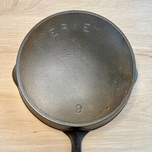 Puede incluir: Una sartén de hierro fundido de color gris oscuro con un solo mango. La palabra "ERIE" está grabada en la parte superior, y el número "9" también es visible. La sartén es circular y parece estar bien sazonada.