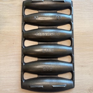 May include: A vintage cast iron muffin pan with six oval molds. The pan is dark gray with the words "No 26", "GRISWOLD", "ERIE", and "958" embossed on it. The pan has handles on each side.