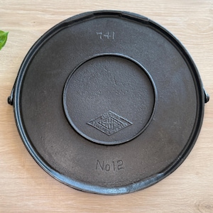 May include: A black cast iron lid with a circular design. The lid has a raised rim and a smaller circular indentation in the center. The number "741" is stamped at the top, and "No 12" at the bottom. A diamond-shaped logo is in the center.