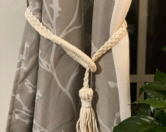 May include: A white cotton rope curtain tieback with a tassel. The tieback is tied around a pair of curtains with a floral pattern.