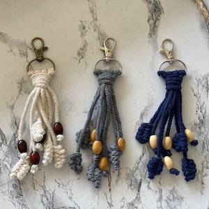 Puede incluir: Tres llaveros de macramé hechos a mano con cuentas de madera. Los llaveros son en tonos blanco, gris y azul.