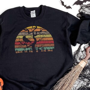 Puede incluir: Sudadera negra con un diseño retro de puesta de sol. El gráfico presenta una silueta de árbol, cuervo y cementerio en tonos naranja, amarillo, verde y marrón. Una prenda con temática de Halloween.