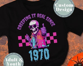 May include: Black t-shirt with a colorful skeleton graphic wearing sunglasses and giving a thumbs up. The text reads "CREEPING IT REAL SINCE 1970". The design includes a checkered pattern and is surrounded by Halloween decorations.