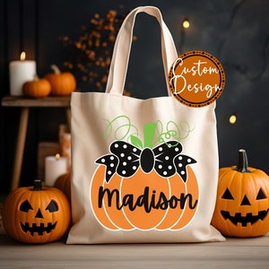 Op de afbeelding: Cr&egrave;me canvas draagtas met een oranje pompoenontwerp. De pompoen heeft een zwarte strik met witte stippen en de naam "Madison" in zwarte letters. Twee uitgesneden pompoenen flankeren de tas, wat een Halloween-thema cre&euml;ert.