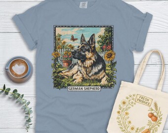 Puede incluir: Camiseta azul claro con un gráfico de un perro pastor alemán en un jardín con girasoles y mariposas. El texto "GERMAN SHEPHERD" está debajo. También se ve una bolsa de mano con el texto "FRESH PRODUCE" y una taza de café.