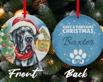 Peut inclure: Une boule de Noël ronde avec un grand danois noir et blanc portant un bonnet de Père Noël sur le devant. Le dos de la boule est bleu avec des flocons de neige blancs et le texte "HAVE A PAWSOME CHRISTMAS Baxter 2024".