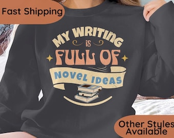 Puede incluir: Sudadera gris oscuro con el texto "My Writing is Full of Novel Ideas" en un estilo retro. El diseño incluye una pila de libros y detalles de estrellas. Una sudadera cómoda e informal.