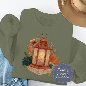 Puede incluir: Sudadera verde oliva con una ilustración detallada de una linterna vintage con detalles florales en naranja, marrón y verde. La linterna tiene un ribete de encaje. También se ve el texto "Luxury T-shirts & Sweatshirts".