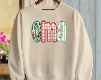 Puede incluir: Una sudadera beige con la palabra "oma" escrita en letras de patchwork de colores. Las letras están delineadas en rosa y tienen una variedad de patrones, incluyendo cuadros, flores y rayas.