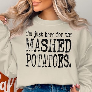 May include: Cream-colored sweatshirt with the text "I'm just here for the MASHED POTATOES." in a distressed black font. The sweatshirt is a casual, long-sleeved garment.