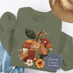 May include: An olive green sweatshirt featuring a patchwork apple design with floral accents. The apple is composed of various fabric patterns and textures, with a stem and leaf. The design includes red, orange, and white flowers. The text "Luxury T-shirts & Sweatshirts" is also visible.